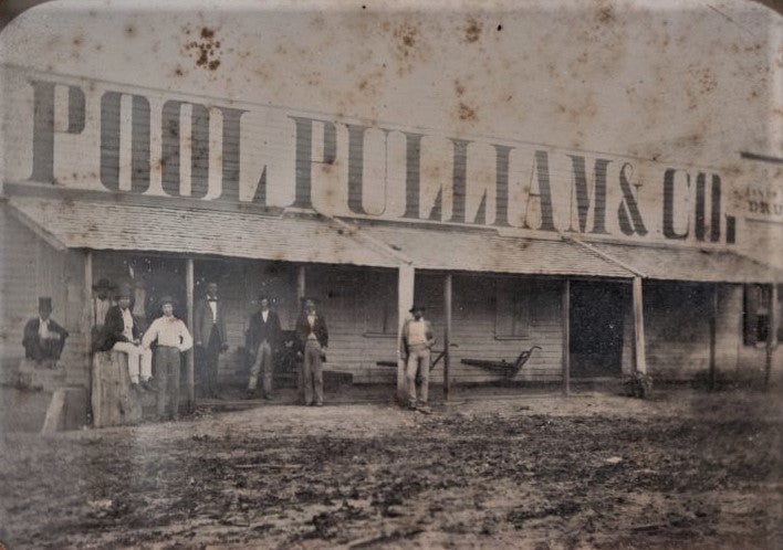 a black-and-white photo of a squat wooden building on the other side of a muddy street